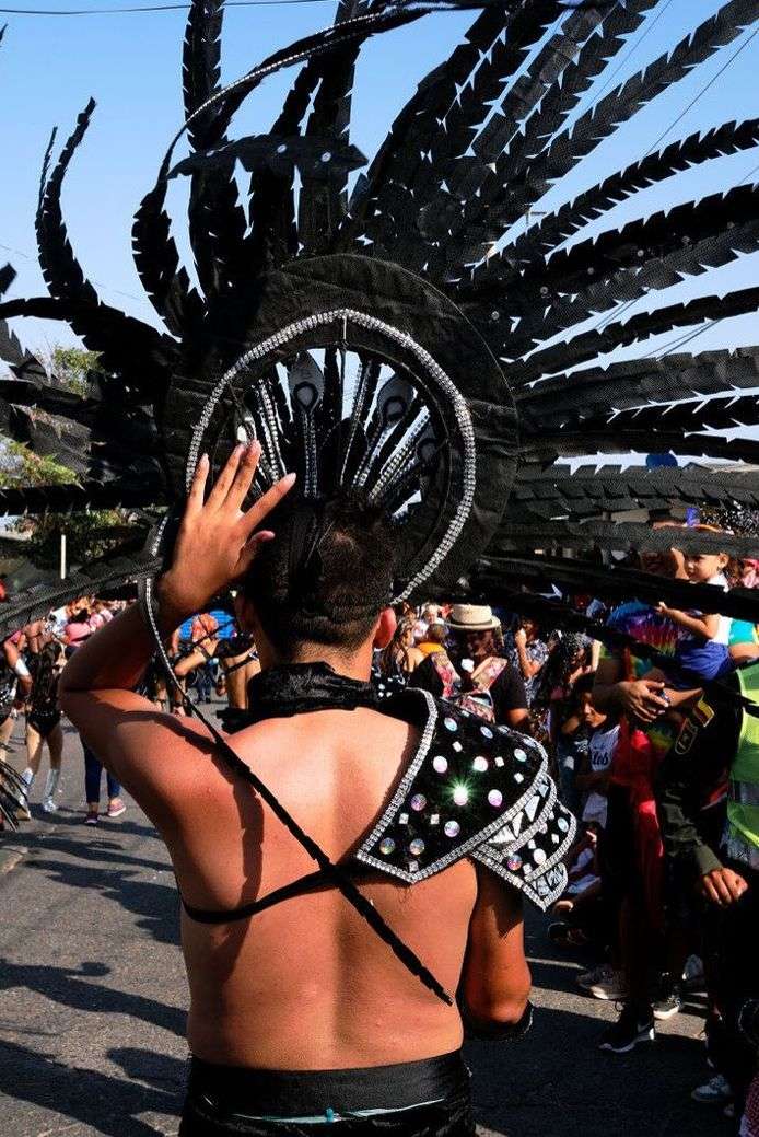A man in a black headdress with feather-like extensions stands outdoors, facing away from the camera, surrounded by a crowd of people.
