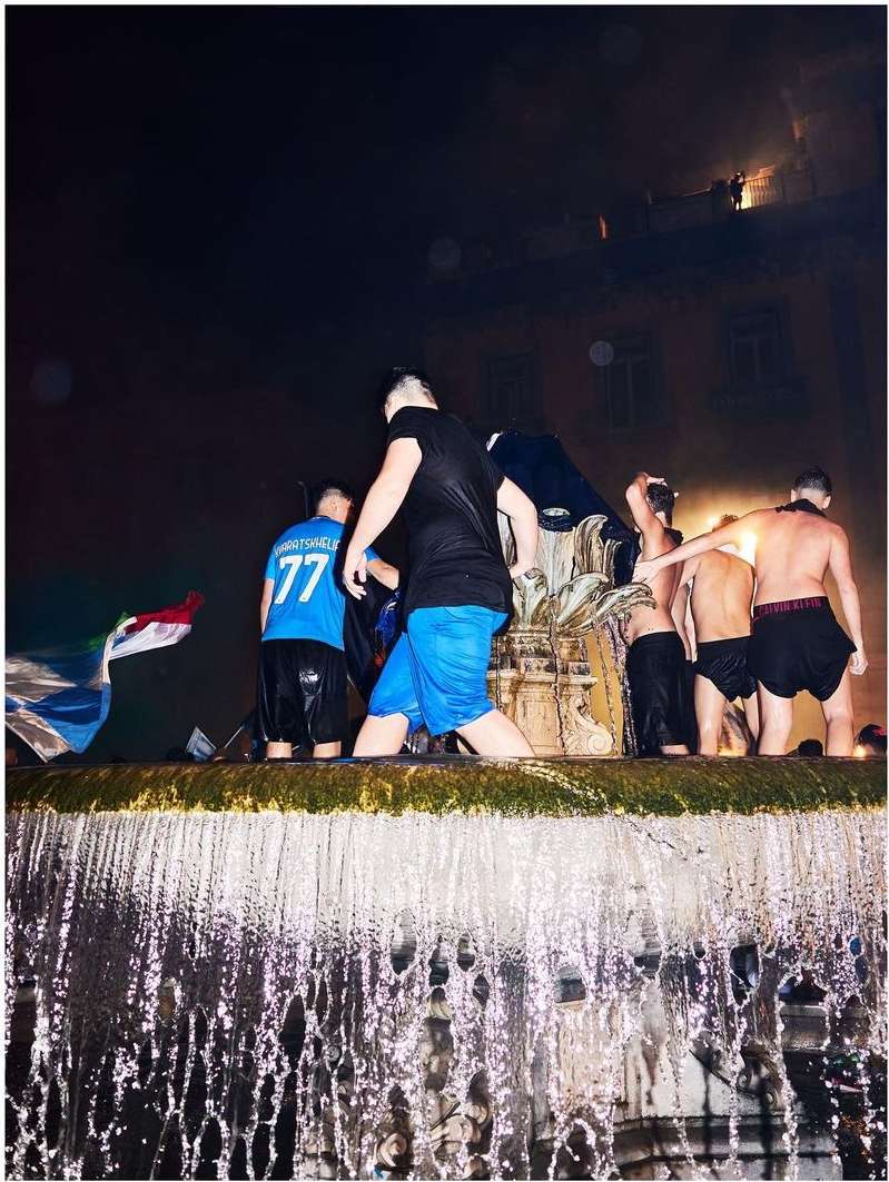 Men celebrate in a fountain at night, captured with a low angle, showing a city building in the background and motion blur from the water.