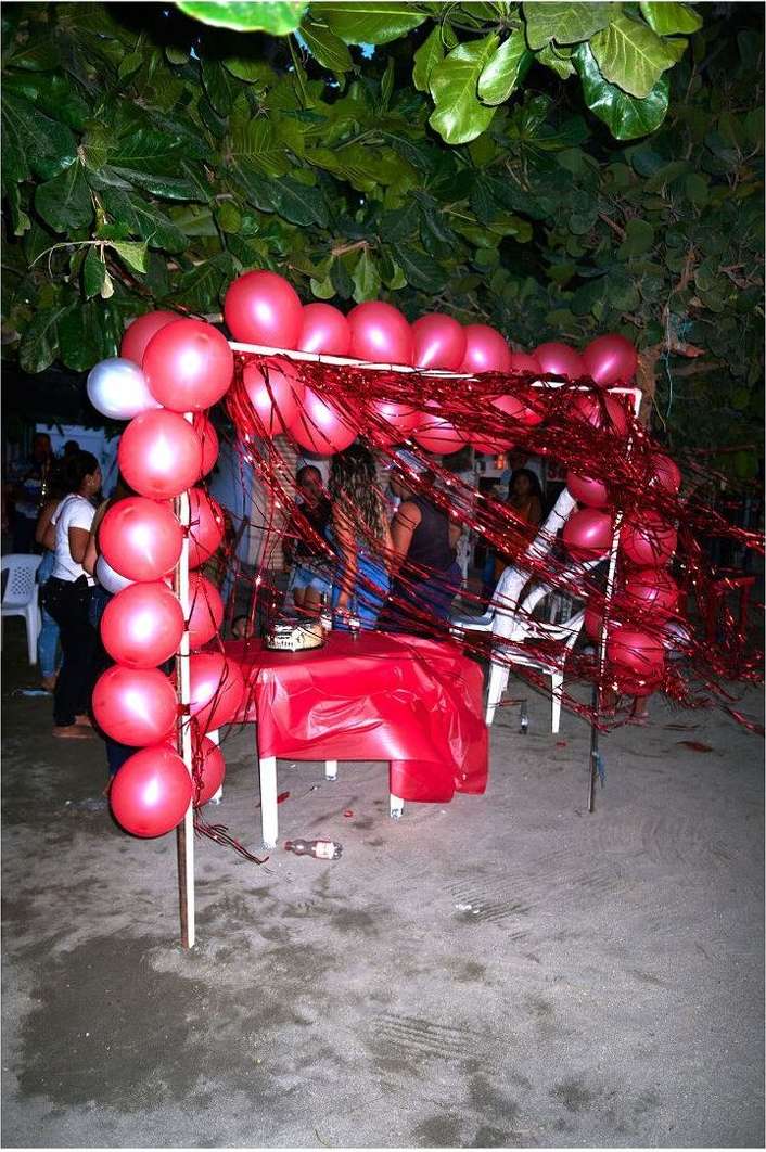 Outdoor party scene at night with a balloon arch, covered table, and several people gathered under trees, creating a festive atmosphere.
