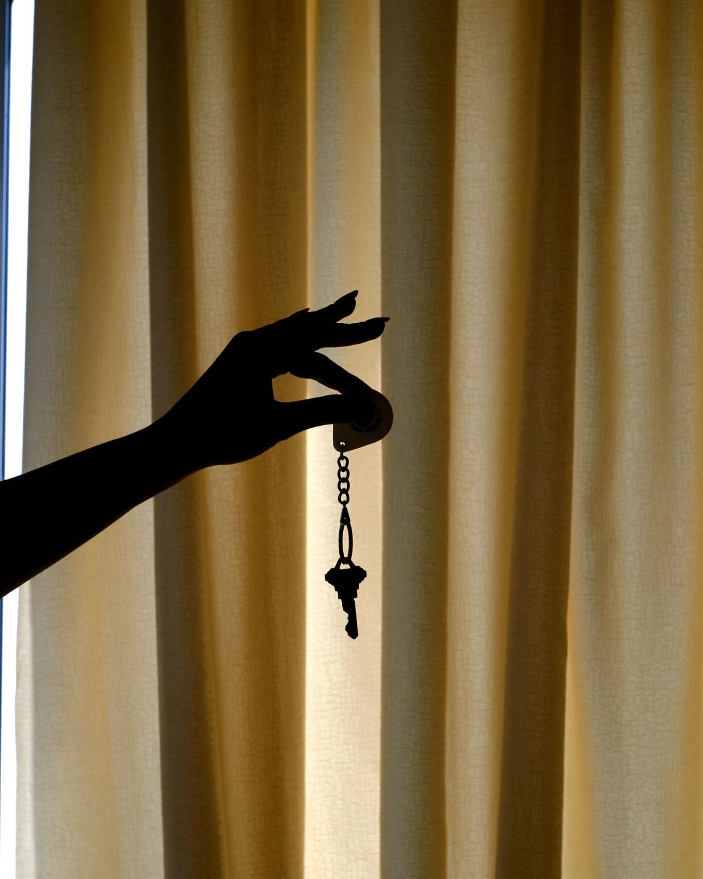 Silhouette of a woman's hand holding keys with a tan curtain backdrop, emphasizing shape and contrast in an indoor setting.