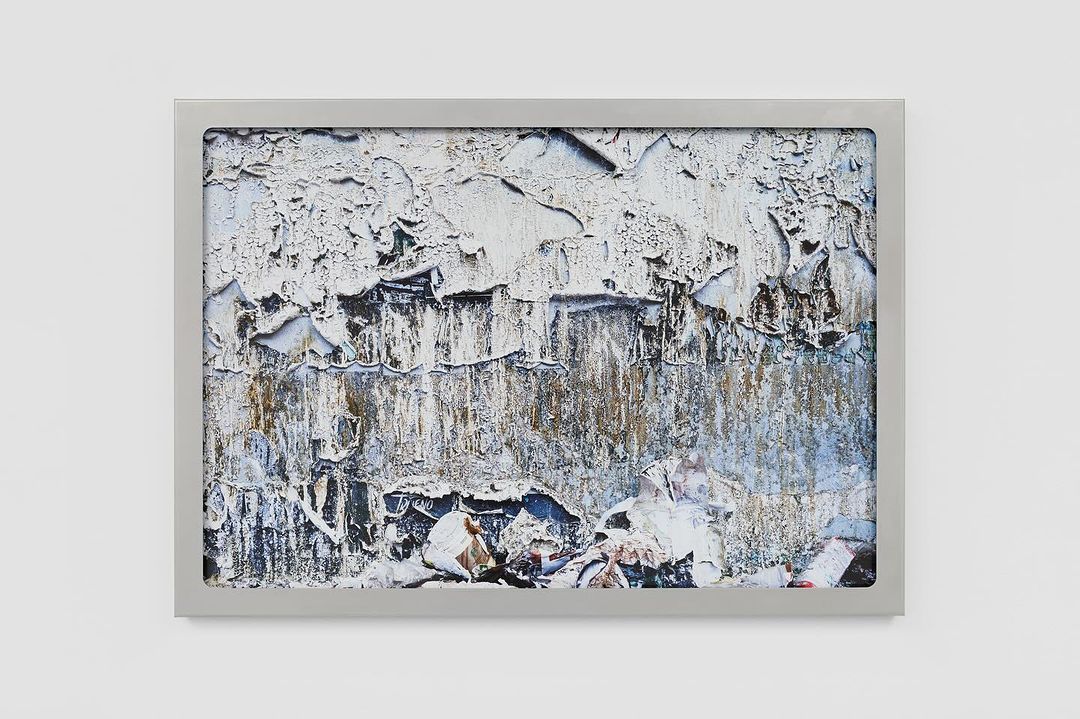 Close-up of a framed abstract art piece with textures, peeling paint, and neutral tones, showcased against a clean white wall.