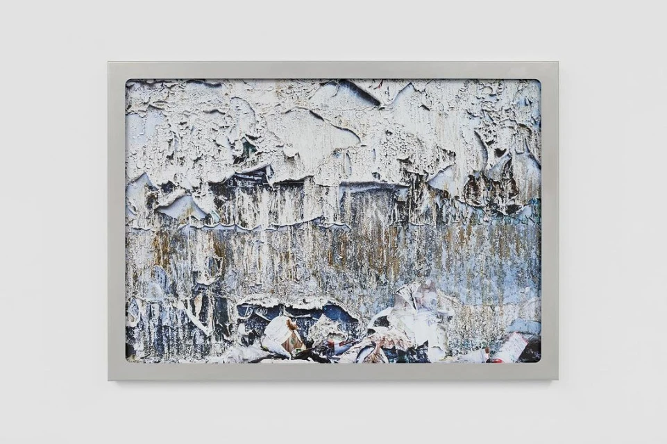 Close-up of a framed abstract art piece with textures, peeling paint, and neutral tones, showcased against a clean white wall.