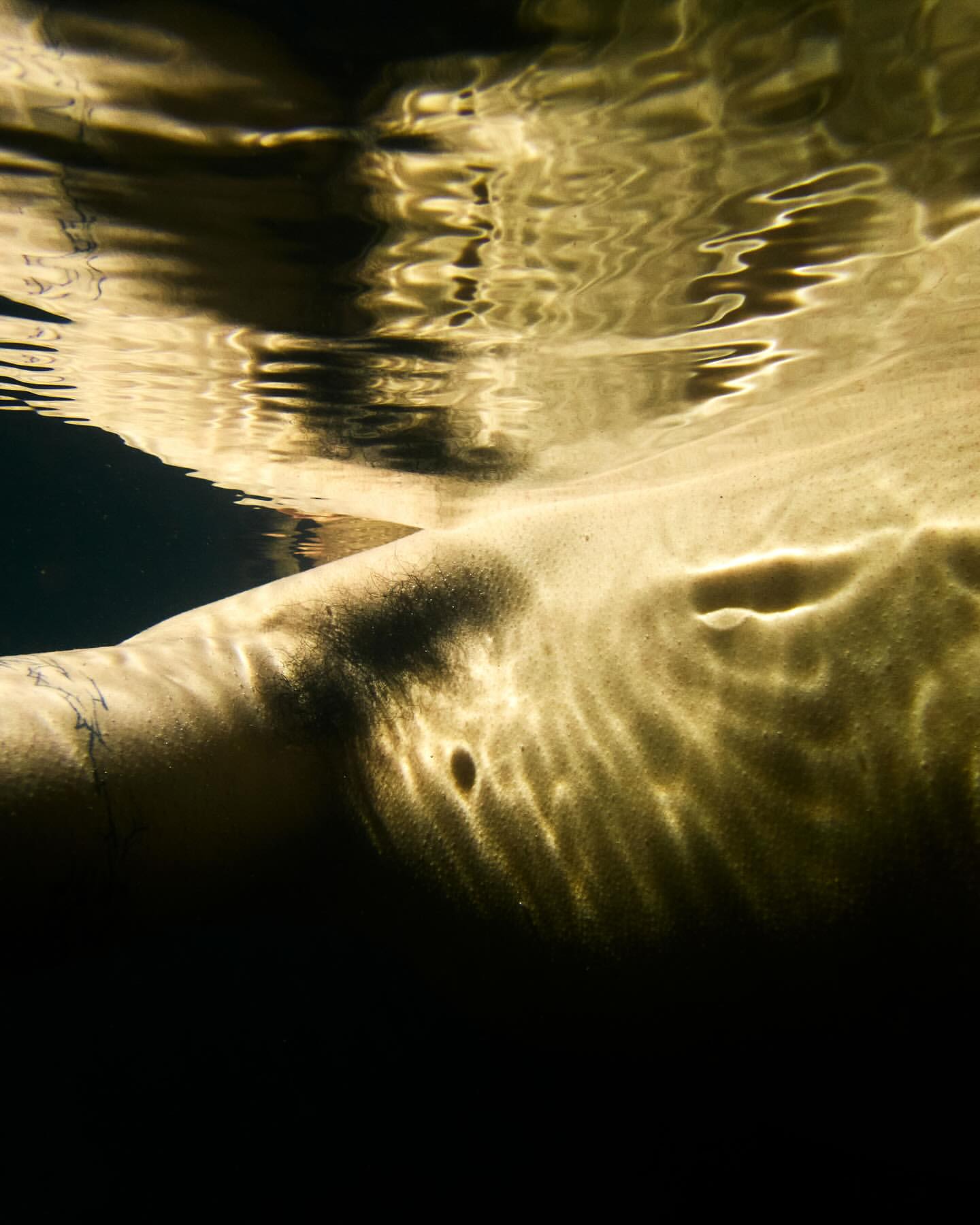 Underwater view shows the side torso of a light-skinned adult illuminated by light filtering through water, against a dark background.