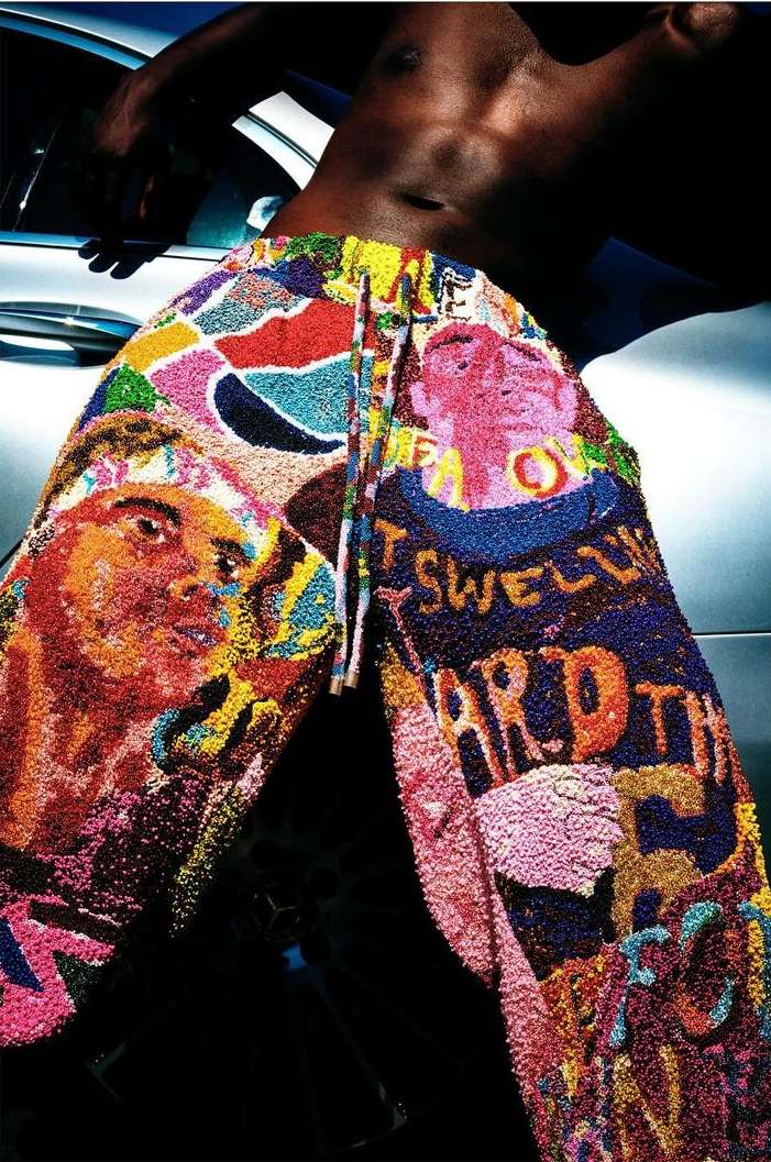 Torso of a Black male in the foreground and his legs are covered in multicolored pants, with a blurred background of the side of a car.
