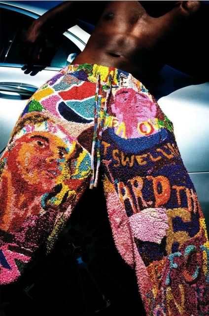 Torso of a Black male in the foreground and his legs are covered in multicolored pants, with a blurred background of the side of a car.
