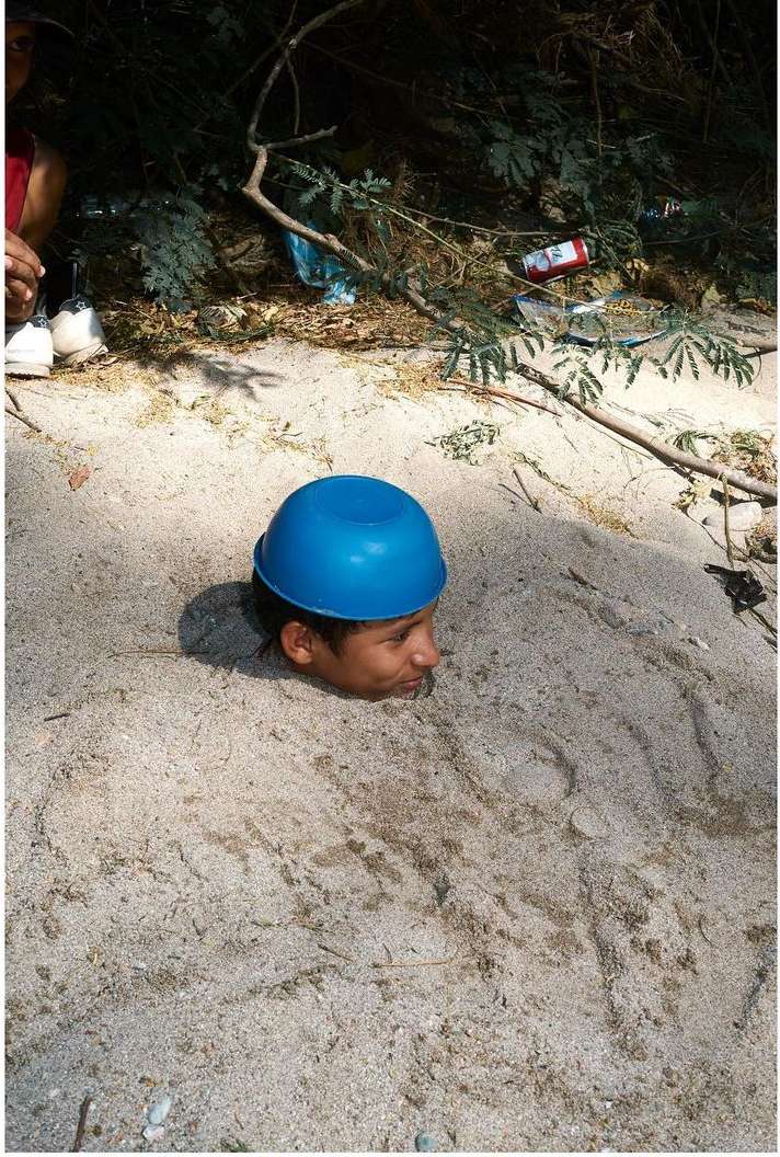 Young person buried in sand up to the neck, wearing a blue bowl on their head, with foliage and trash in the blurred background.