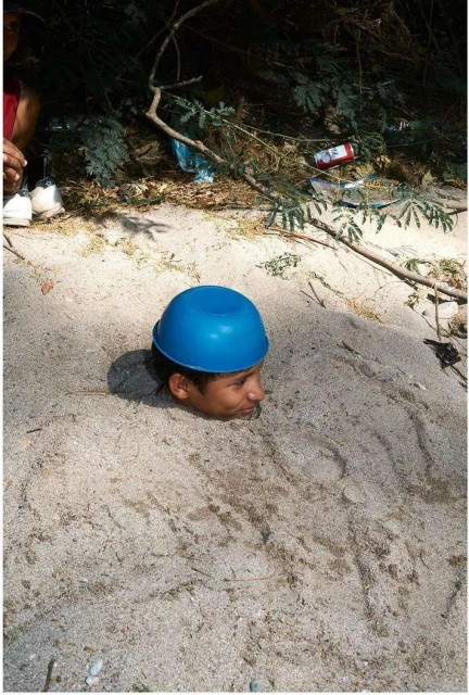 Young person buried in sand up to the neck, wearing a blue bowl on their head, with foliage and trash in the blurred background.