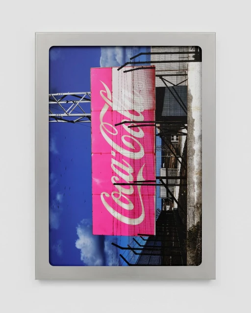 Framed Coca-Cola advertisement partially obscured by a chain link fence against blue sky. A metal framework supports the sign.