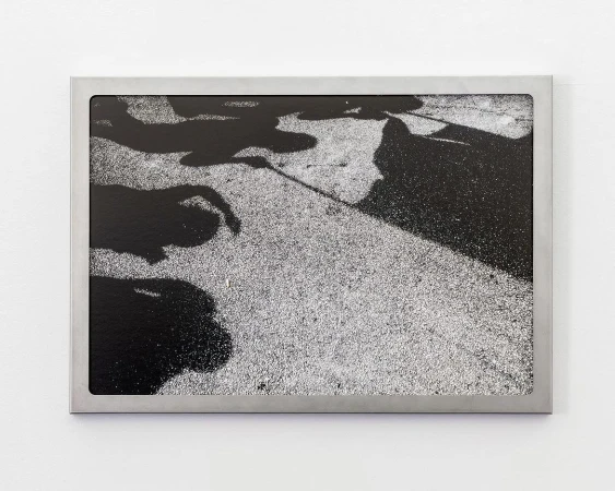 High-angle black-and-white photographic art print in a silver frame, showing dark human shadows on a grainy gray sidewalk.