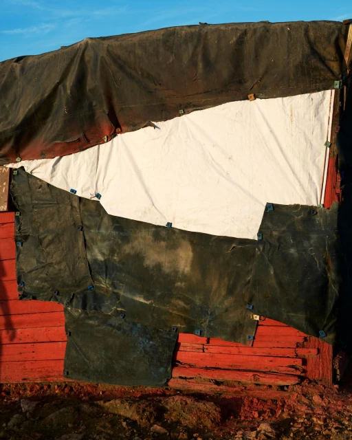 Abstract view of a makeshift structure with patched-up tarps and bright red wooden planks against a clear blue sky.