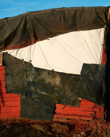 Abstract view of a makeshift structure with patched-up tarps and bright red wooden planks against a clear blue sky.