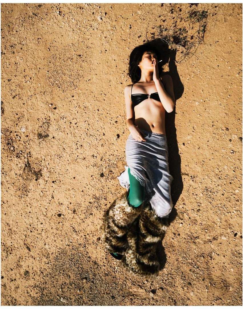 Overhead shot of a woman in lingerie and a skirt lying on a textured ground surface in natural light, with a fur-like boot cover.