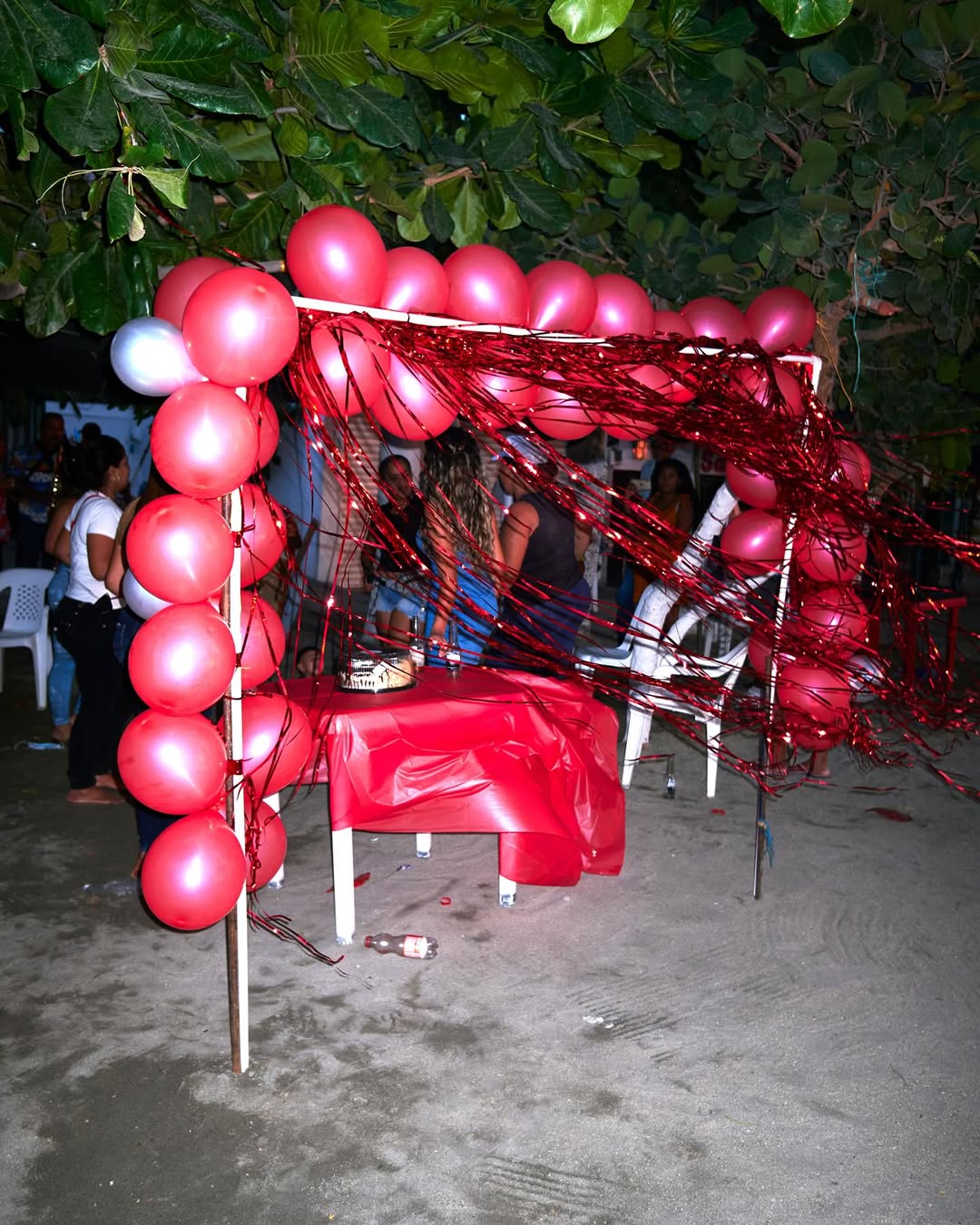 Outdoor party scene with red and white balloons forming an archway, people gathered in the background, set on a sandy surface near trees.