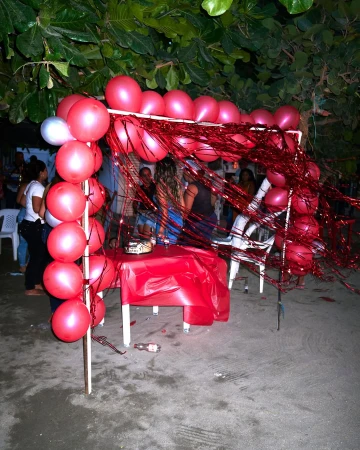 Outdoor party scene with red and white balloons forming an archway, people gathered in the background, set on a sandy surface near trees.
