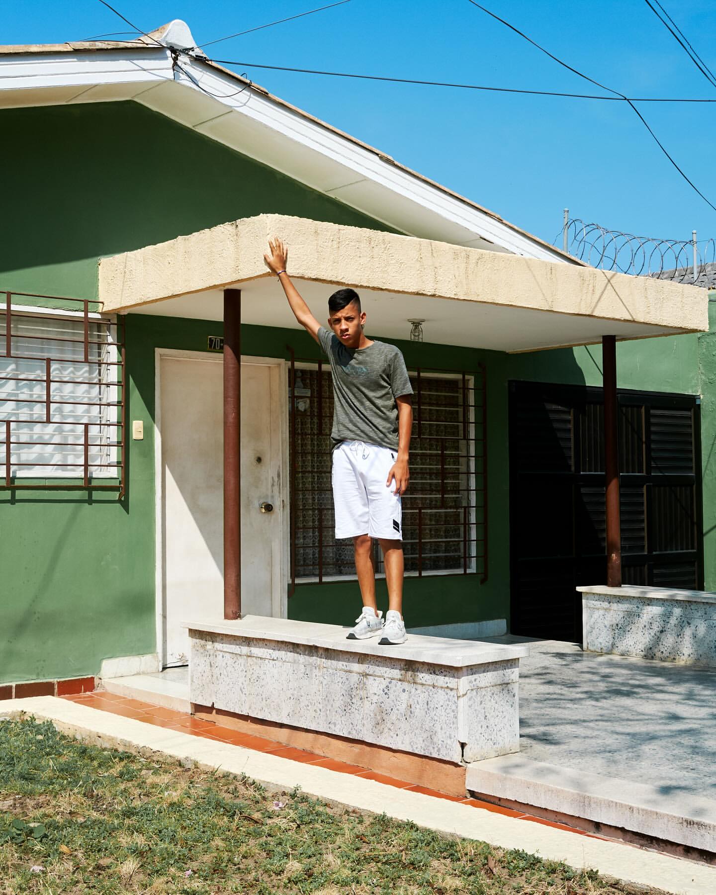 A young man stands on a low wall in front of a green house on a sunny day, raising his arm up to the roof of the porch.