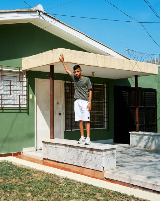 A young man stands on a low wall in front of a green house on a sunny day, raising his arm up to the roof of the porch.