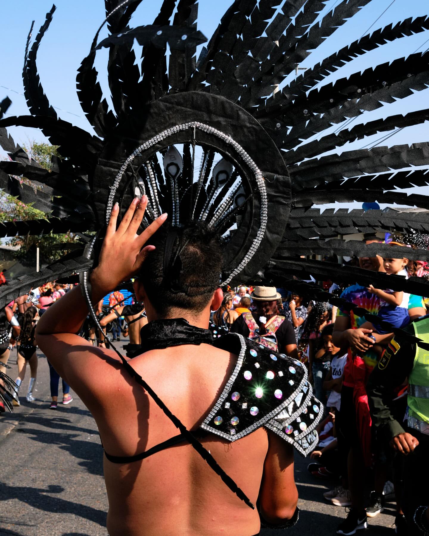 A man in an elaborate black feathered headdress and decorative shoulder piece stands in a parade or outdoor celebration on a sunny day.