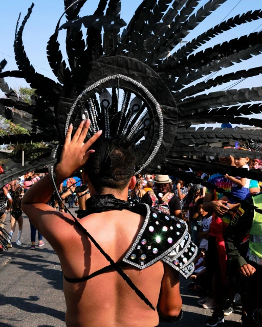 A man in an elaborate black feathered headdress and decorative shoulder piece stands in a parade or outdoor celebration on a sunny day.