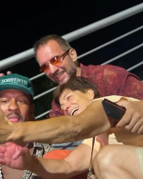 Three adults pose cheek-to-cheek, smiling and laughing in an evening setting, one wearing a 'Sorrento' cap and red tinted glasses.