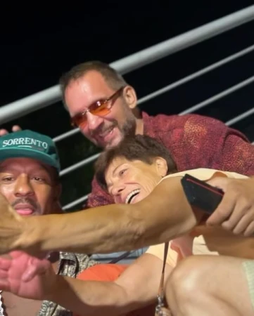 Three adults pose cheek-to-cheek, smiling and laughing in an evening setting, one wearing a 'Sorrento' cap and red tinted glasses.