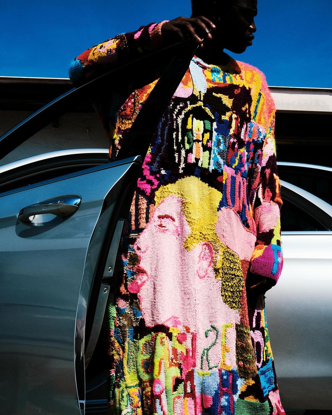Dark-skinned man in bright patchwork sweater posing next to a silver car against a blue sky. The sweater has a face on it.