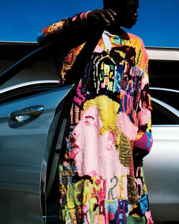 Dark-skinned man in bright patchwork sweater posing next to a silver car against a blue sky. The sweater has a face on it.