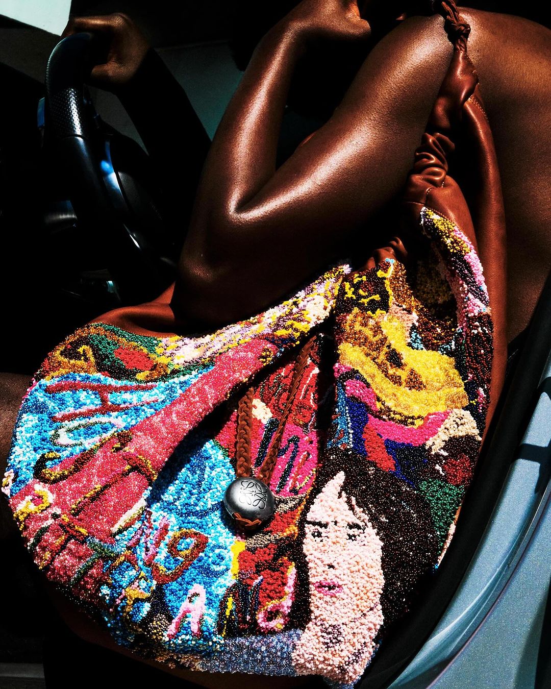 Close-up shot of a woman's arm and shoulder holding a colorful beaded handbag in a car; the bag features a portrait design.