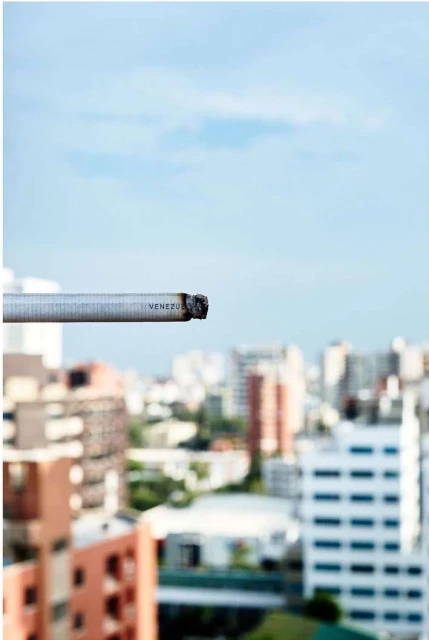 Lit cigarette in focus with a blurred city skyline in the background on a sunny day with blue sky and scattered clouds.