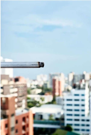 Lit cigarette in focus with a blurred city skyline in the background on a sunny day with blue sky and scattered clouds.