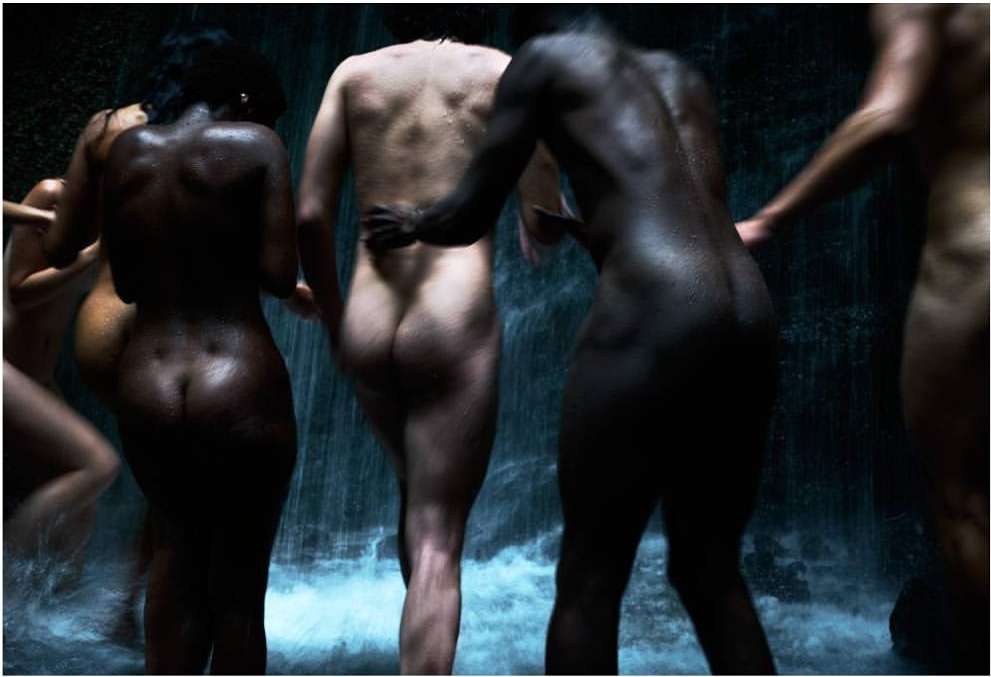 A group of nude adults of varying ethnicities are gathered together in front of a small waterfall, possibly for a fine art photo.