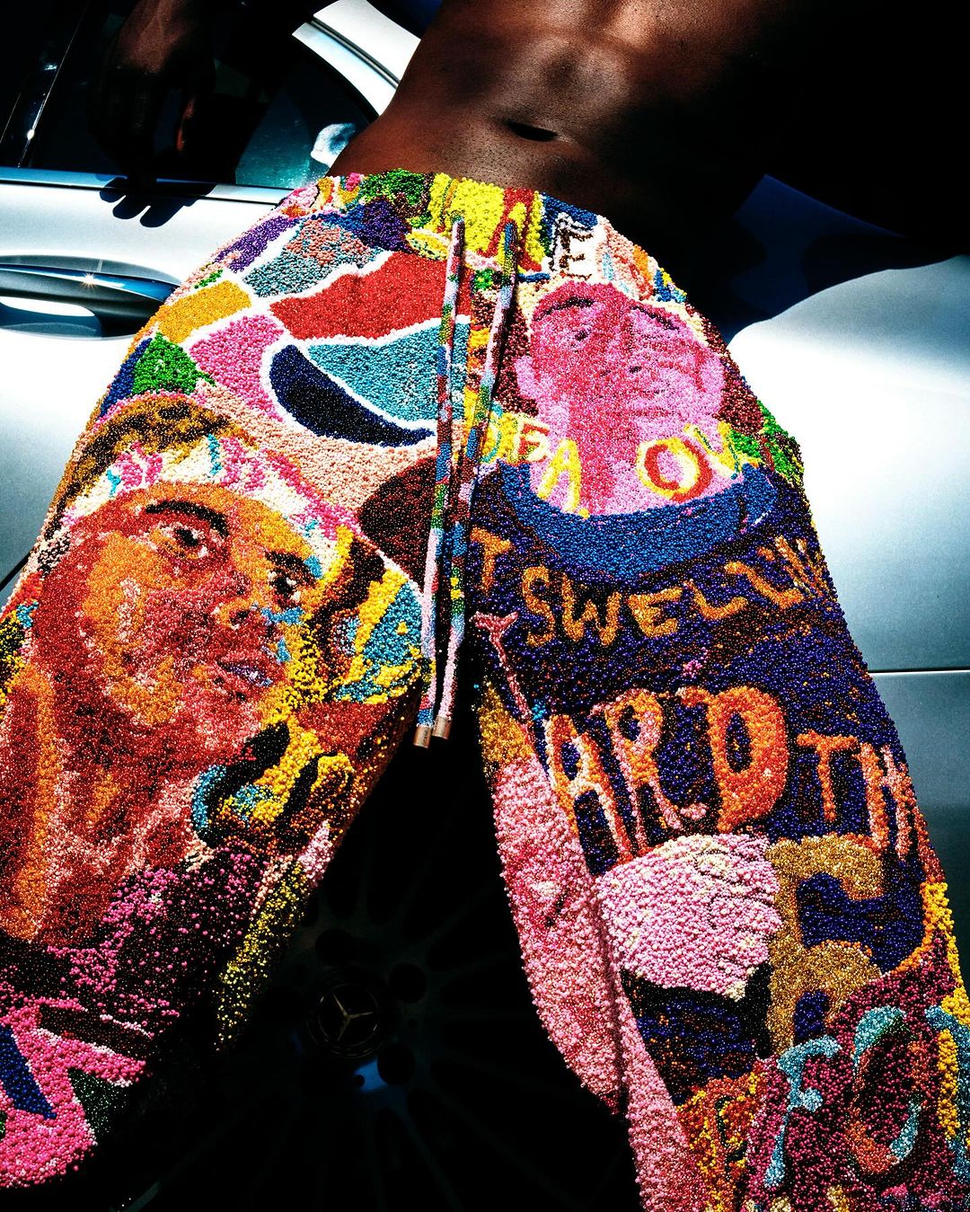 Close-up portrait of a shirtless man wearing brightly colored graphic patterned textured trousers leaning against a car.