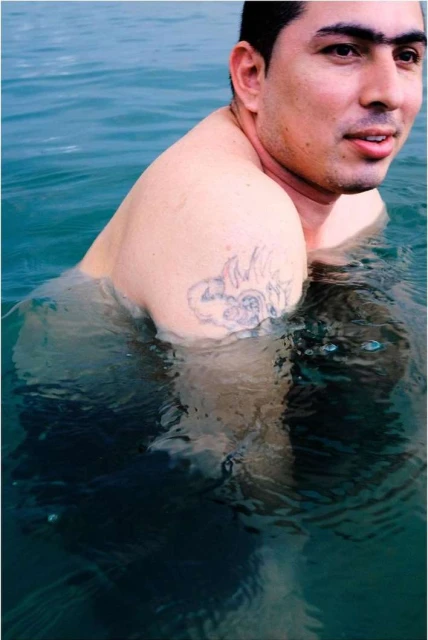 Young man stands waist-deep in turquoise water with a shoulder tattoo and a slight smile, enjoying a sunny day at the beach.