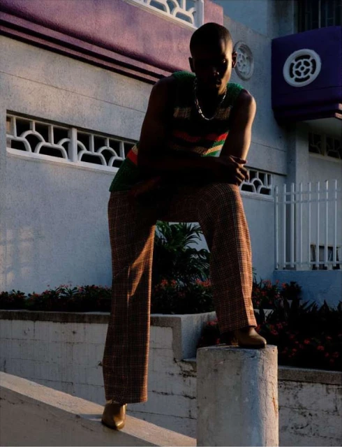 Man in vintage-style outfit posing outdoors against building with brown plaid pants, green top and loafers in golden hour light.