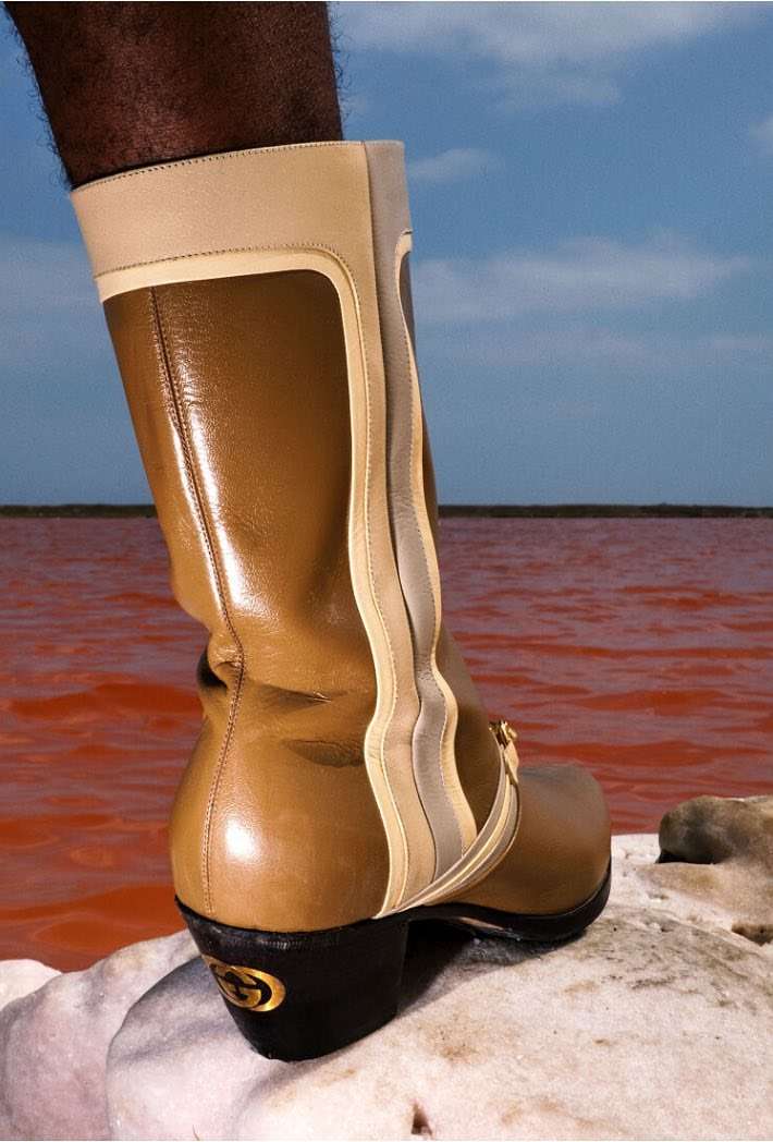 Tan leather knee-high boot with light beige accents, set on a light stone with a red body of water and a light blue sky backdrop.