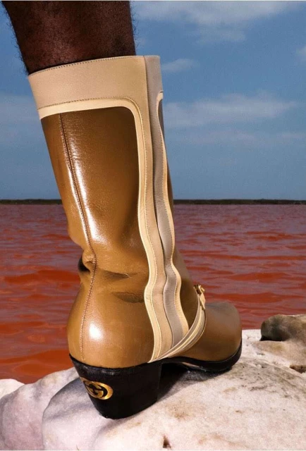 Tan leather knee-high boot with light beige accents, set on a light stone with a red body of water and a light blue sky backdrop.