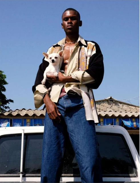 Adult male poses outdoors in an open shirt, jeans, and necklace while holding a white chihuahua in arms on the back of a white truck.