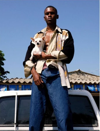 Adult male poses outdoors in an open shirt, jeans, and necklace while holding a white chihuahua in arms on the back of a white truck.