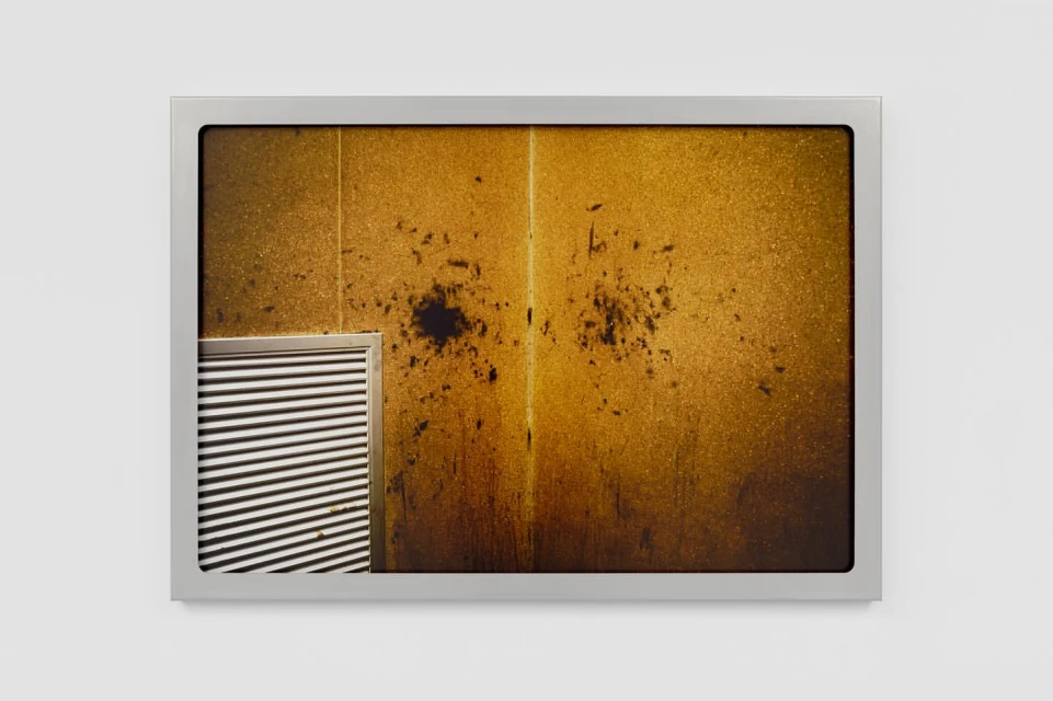 Abstract photograph of a weathered gold surface with dark stains, framed in silver, against a plain white background.
