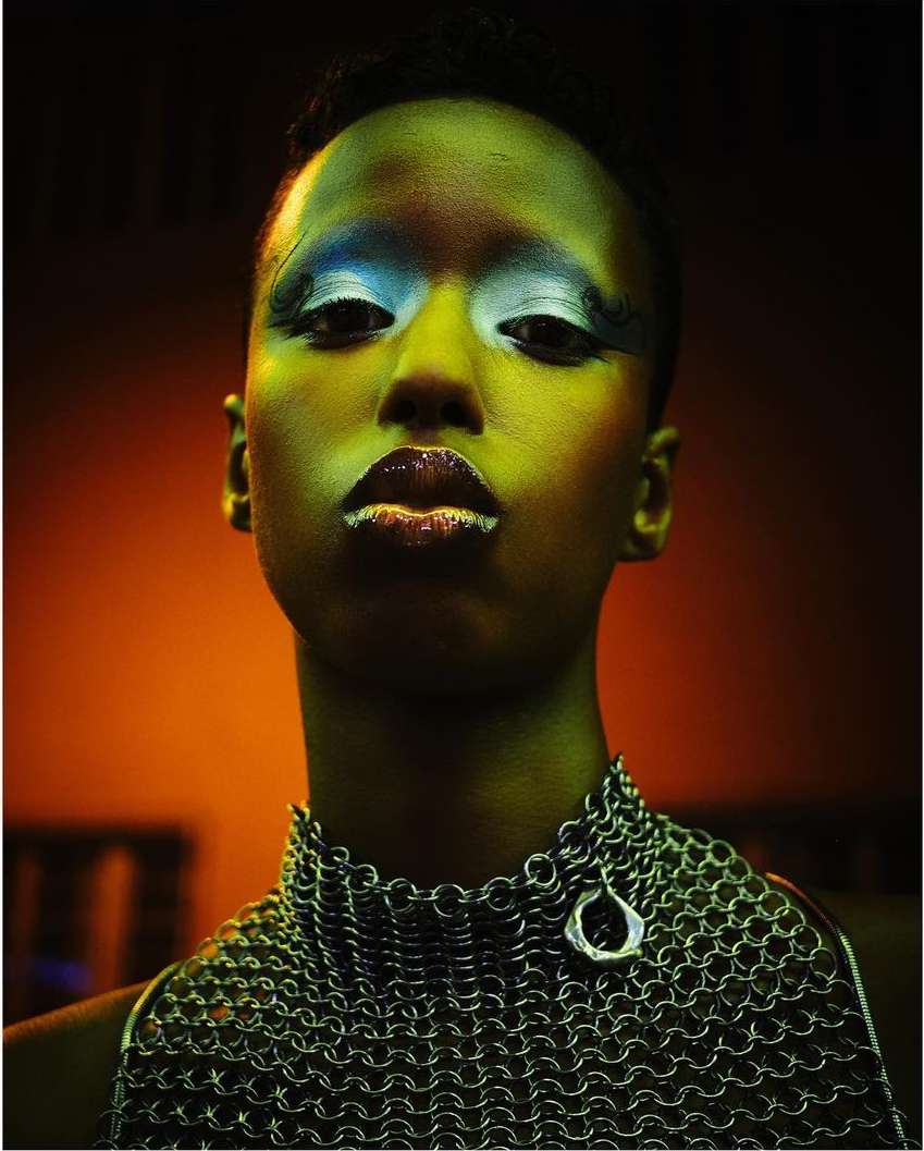 Close-up portrait of a person with dark skin, vibrant eye makeup, and a metal chainmail garment against a dark orange backdrop.