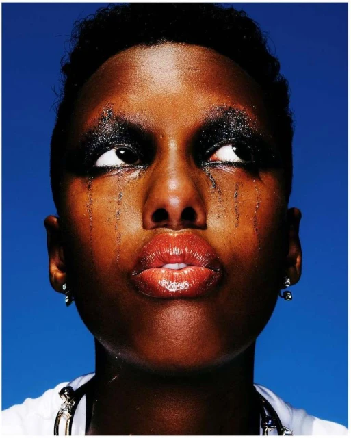 Close-up portrait of a black woman with dramatic black eye makeup, glossy lips, and teardrop accents against a blue background.