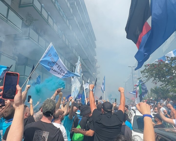 Outdoor celebration with a large group of people holding flags and cell phones on a city street with buildings under a blue sky.