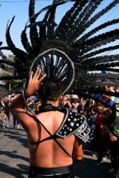 Rear view of a performer wearing a large black feathered headdress and ornate sequined shoulder armor on a street.