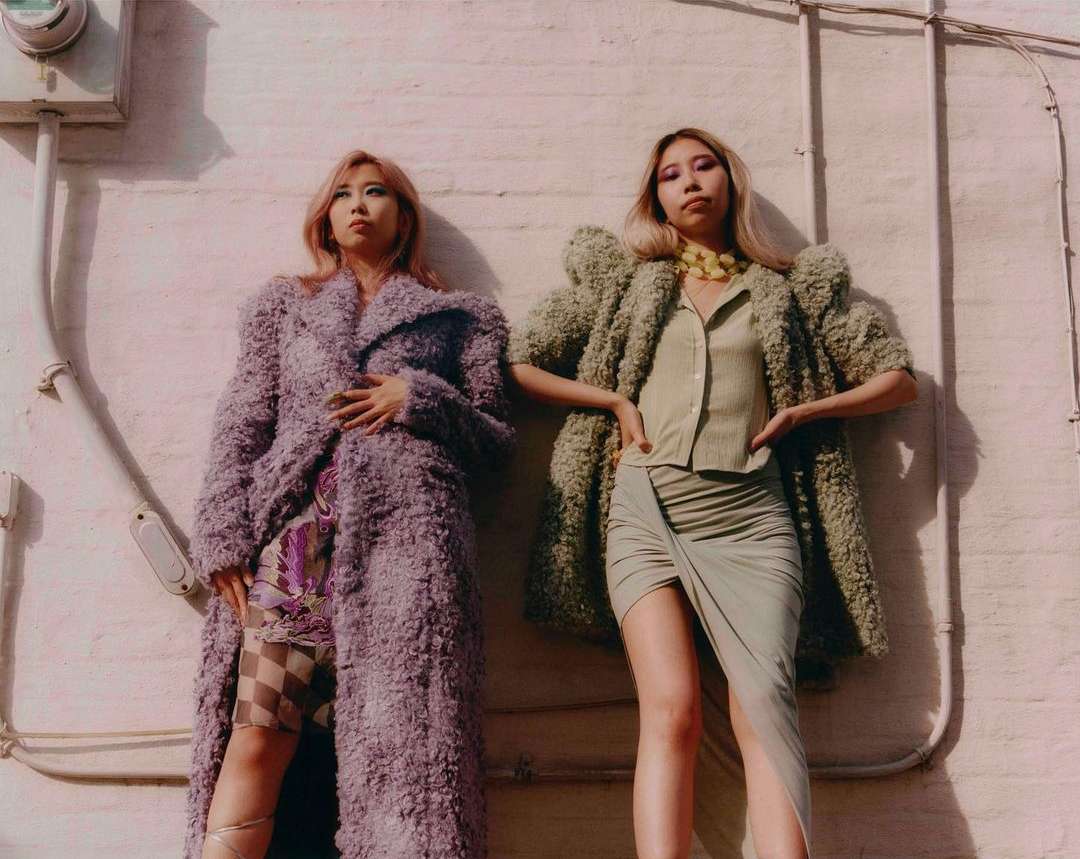 Two women stand side-by-side outside, posing in oversized, textured coats, with sunlight casting shadows on the pale pink and white wall behind them.