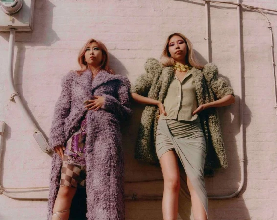 Two women stand side-by-side outside, posing in oversized, textured coats, with sunlight casting shadows on the pale pink and white wall behind them.