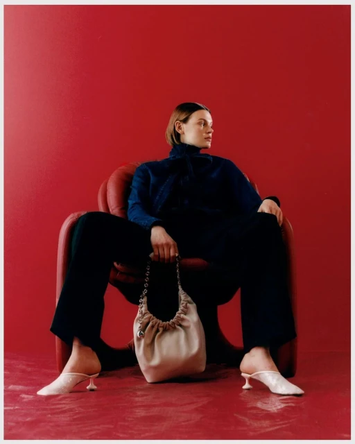 A woman in a dark turtleneck sweater and trousers sits in a red chair holding a light-colored handbag, with a red backdrop.