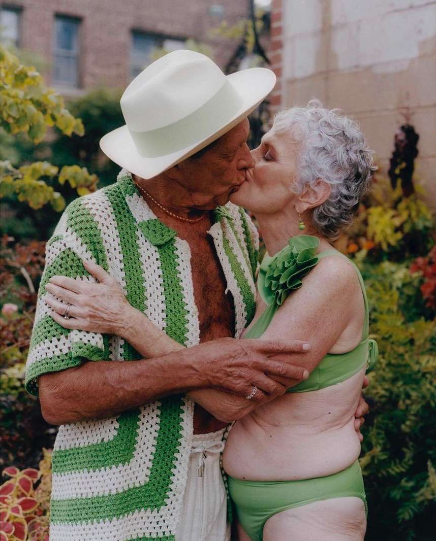 Elderly couple kissing outdoors in green swimsuits and crocheted clothing amidst lush greenery, capturing a moment of affection.