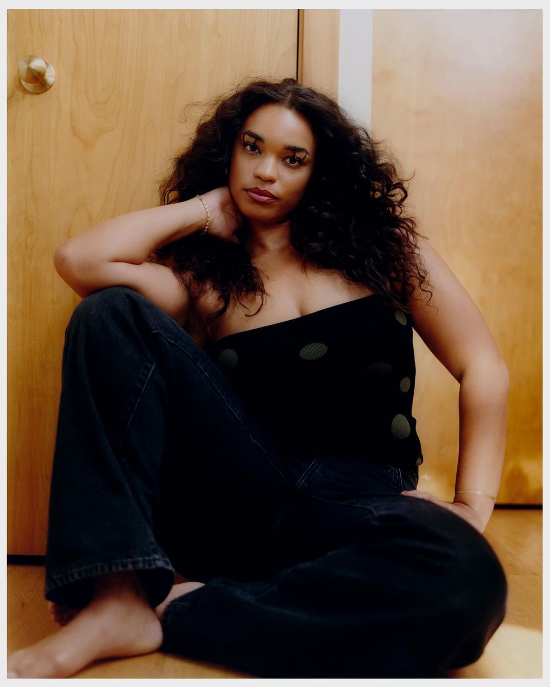 Portrait of an adult woman with long, curly hair. She is wearing a black top and jeans, sitting in front of a light brown door.