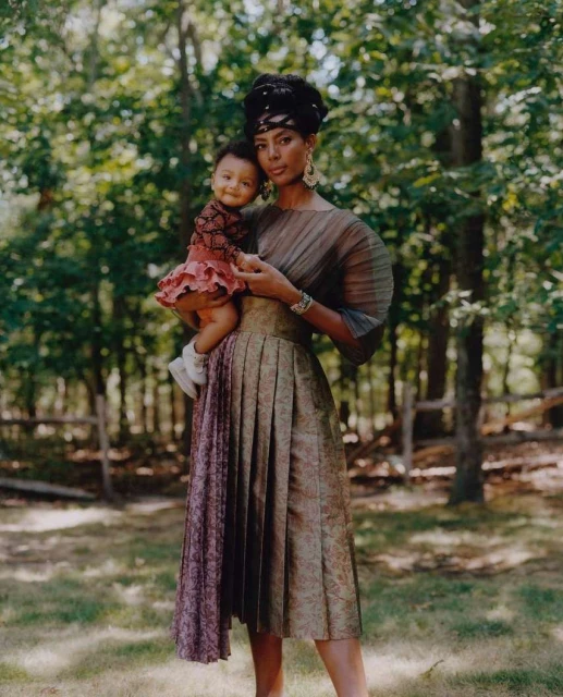 A stylish mother holds her young child in a verdant outdoor setting, both elegantly dressed and accessorized, bathed in soft, natural light.
