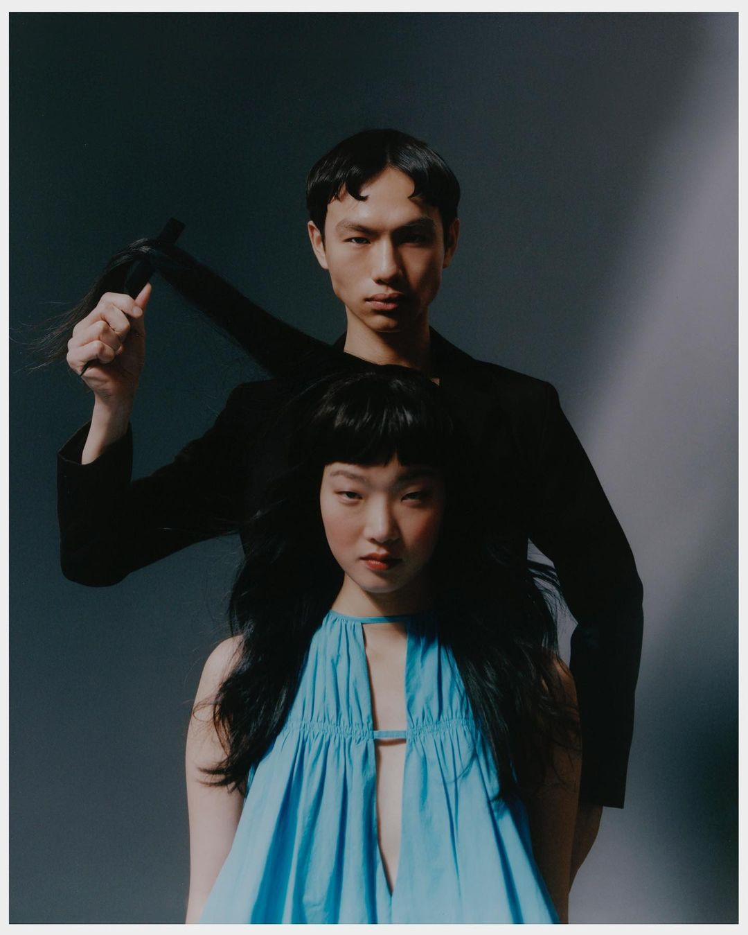 Fashion shot with a stylish couple; the man in a blazer holds a braid of black hair above the woman in a blue dress, against a grey background.