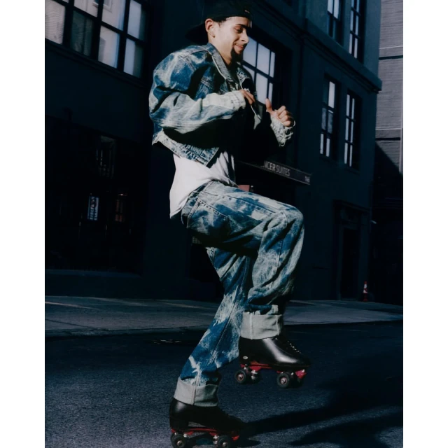 Young adult roller skating on a city street at night, wearing a baseball cap, denim jacket and jeans, captured in an eye-level shot.